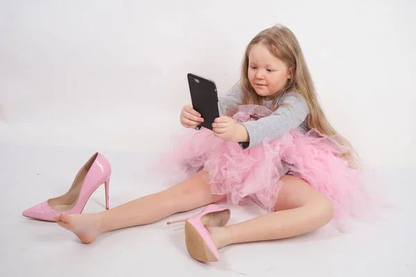 小さな白人の子供の女の子が座って 母のハイヒールのピンクの靴を置き 白いスタジオの背景に彼女のスマートフォンでそれをすべてと Selfies の写真を撮る — ストック写真