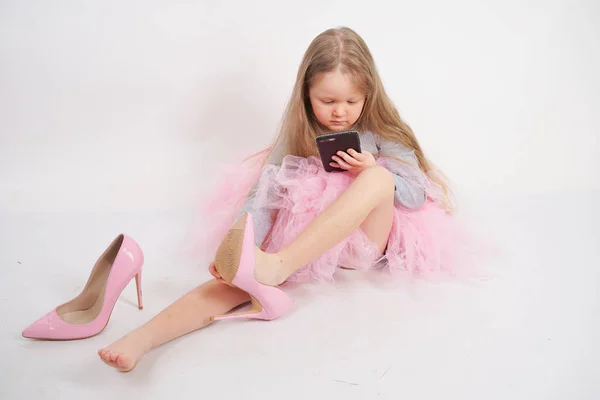 小さな白人の子供の女の子が座って 母のハイヒールのピンクの靴を置き 白いスタジオの背景に彼女のスマートフォンでそれをすべてと Selfies の写真を撮る — ストック写真