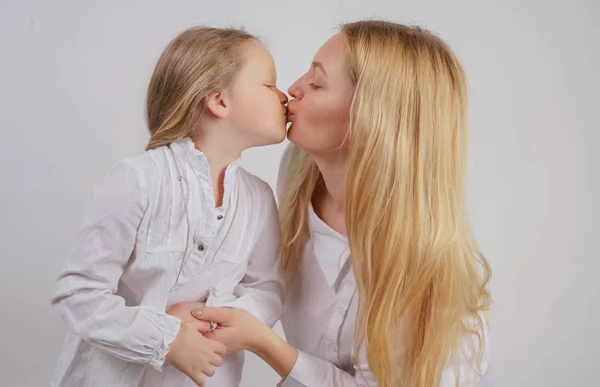 mom and daughter in white shirts with long blonde hair posing on a solid background in the Studio. charming family takes care of each other and hugs.