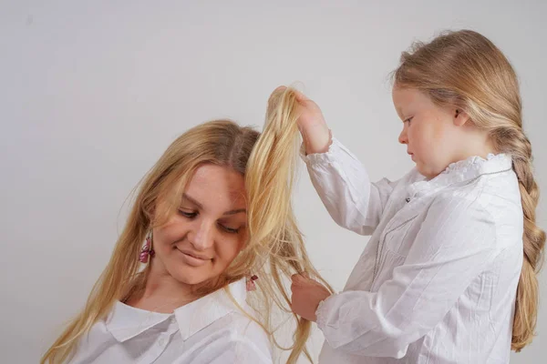 mom and daughter in white shirts with long blonde hair posing on a solid background in the Studio. a charming family takes care of each other and makes braids.