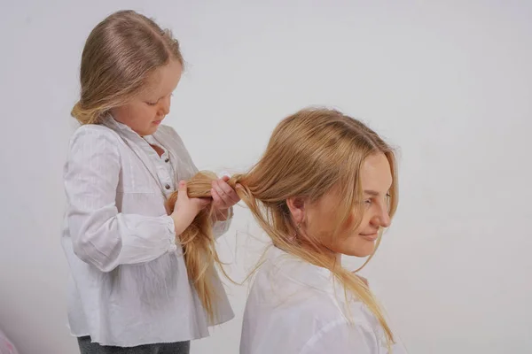 スタジオでの固体の背景にポーズ長いブロンドの髪と白いシャツでママと娘 魅力的な家族はお互いの世話をし 三つ編みになります — ストック写真