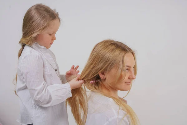スタジオでの固体の背景にポーズ長いブロンドの髪と白いシャツでママと娘 魅力的な家族はお互いの世話をし 三つ編みになります — ストック写真