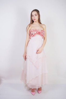 büyüleyici bir genç beyaz kız göğsünde çiçek yaprakları ile pembe uzun balo elbisesi duruyor ve stüdyo beyaz bir arka planda pozlar