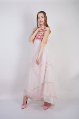 büyüleyici bir genç beyaz kız göğsünde çiçek yaprakları ile pembe uzun balo elbisesi duruyor ve stüdyo beyaz bir arka planda pozlar