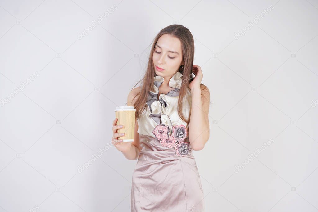 cute teen caucasian girl in a pretty evening dress gives a paper Cup of coffee, treating the interlocutor and smiling, on a white background in the Studio