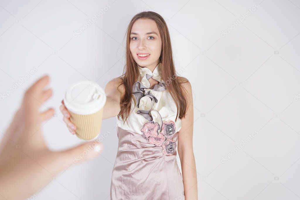 cute teen caucasian girl in a pretty evening dress gives a paper Cup of coffee, treating the interlocutor and smiling, on a white background in the Studio