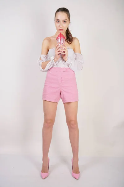 sweet girl in transparent blouse, summer funny plaid shorts and pink high heels posing with a glass cup of water on white background in studio