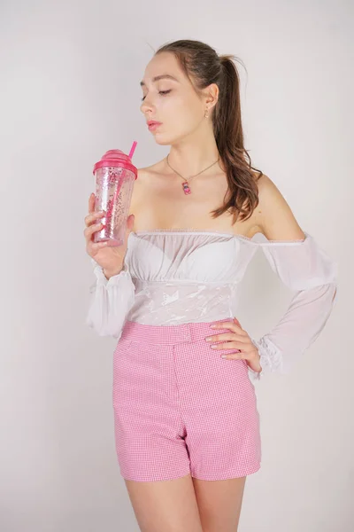 sweet girl in transparent blouse, summer funny plaid shorts and pink high heels posing with a glass cup of water on white background in studio