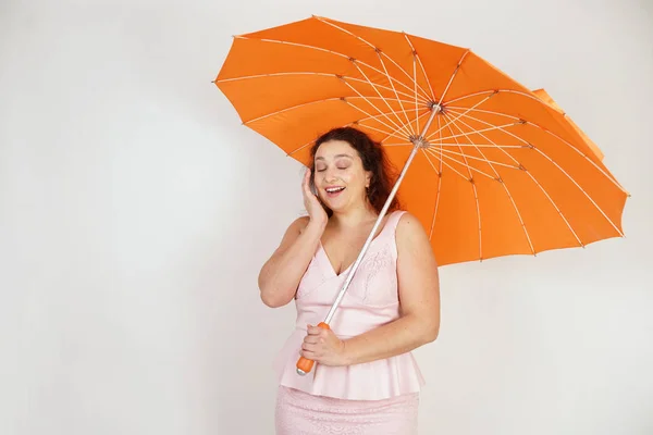 feminine woman with plus size body in pink dress with orange big heart shaped umbrella posing on white background in Studio