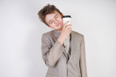 beyaz stüdyo arka plan üzerinde elinde kahve kağıt Fincan ile gri iş elbisesi içinde yorgun genç çocuk