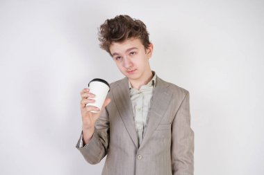 beyaz stüdyo arka plan üzerinde elinde kahve kağıt Fincan ile gri iş elbisesi içinde yorgun genç çocuk