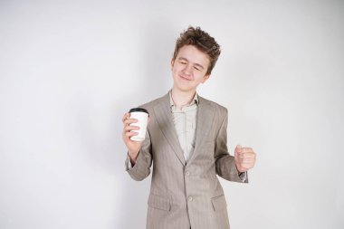 Elinde bir bardak içki olan beyaz takım elbiseli genç beyaz adam, stüdyodaki beyaz bir arkaplanda elde ettiği başarıdan memnuniyet duyuyor.