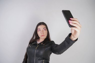 kaya tarzı siyah deri ceket büyüleyici esmer Studio beyaz bir arka plan üzerinde bir akıllı telefon ile duruyor