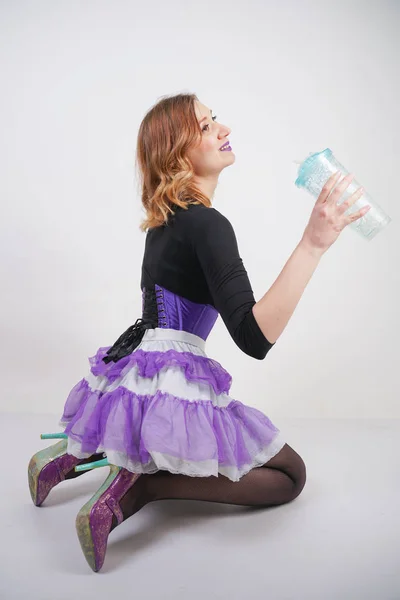 pretty slender girl in purple corset with tutu skirt and mesh black pantyhose standing with blue glass of water on white studio background