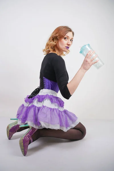 pretty slender girl in purple corset with tutu skirt and mesh black pantyhose standing with blue glass of water on white studio background
