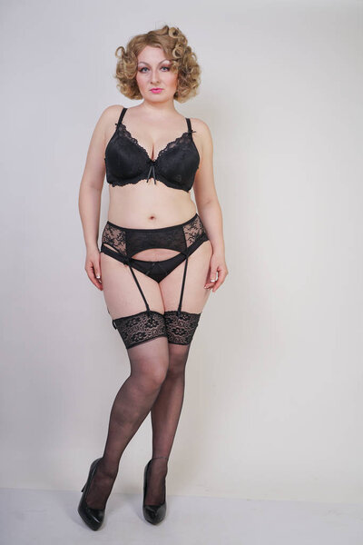 pretty caucasian chubby woman with plus size body and pale skin wearing black sexy underwear with stockings and garterbelt on white studio background