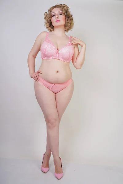 Pretty caucasian chubby woman with plus size body and pale skin wearing pink lace sexy underwear on white studio background isolated