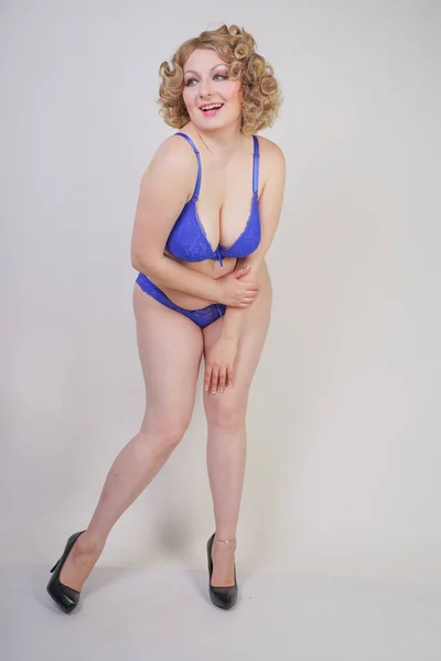 Pretty caucasian chubby woman with plus size body and pale skin wearing blue lace sexy underwear and high heels on white studio background