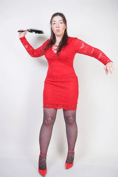 angry negative woman with a chubby body stands in a red dress and holds a black skillet on white studio background. the girl is angry and dissatisfied with the diet and her late coming husband.