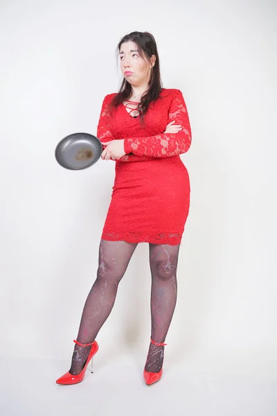 angry negative woman with a chubby body stands in a red dress and holds a black skillet on white studio background. the girl is angry and dissatisfied with the diet and her late coming husband.