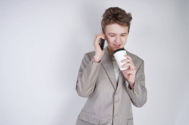 gri bir iş elbisesi içinde kafkas genç stüdyoda beyaz bir arka plan üzerinde bir cep telefonu ve bir kağıt Fincan kahve ile duruyor