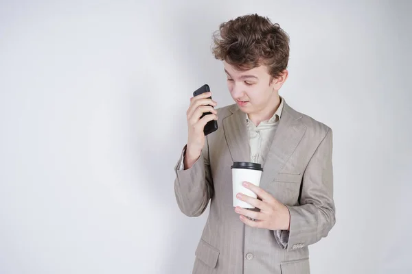 unhappy angry teen in grey business suit stands with mobile phone and paper Cup of coffee on white background in Studio
