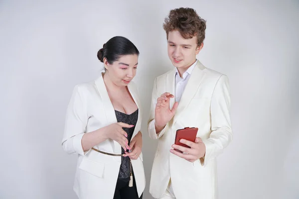 白いビジネススーツを着た白人のティーンエイジャーは フレンドリーな大人のアジアの女性の隣に立って 白い背景に携帯電話を保持しています — ストック写真