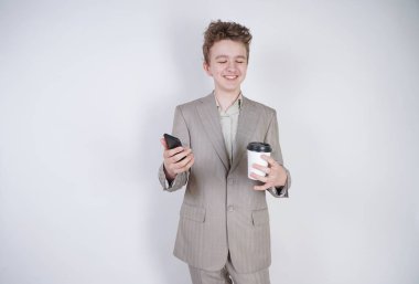 komik gülümseyen genç elinde akıllı telefon ile eğleniyor. beyaz stüdyo arka plan üzerinde kahve kağıt fincan ile ayakta gri iş elbisesi giyen genç çocuk.