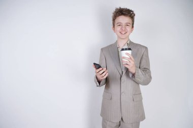 komik gülümseyen genç elinde akıllı telefon ile eğleniyor. beyaz stüdyo arka plan üzerinde kahve kağıt fincan ile ayakta gri iş elbisesi giyen genç çocuk.