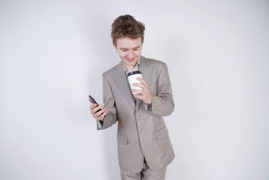 komik gülümseyen genç elinde akıllı telefon ile eğleniyor. beyaz stüdyo arka plan üzerinde kahve kağıt fincan ile ayakta gri iş elbisesi giyen genç çocuk.