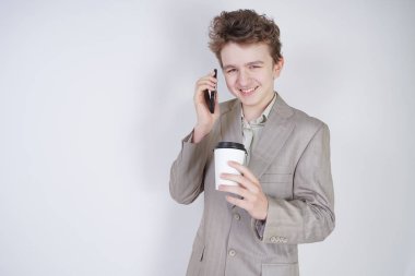 komik gülümseyen genç elinde akıllı telefon ile eğleniyor. beyaz stüdyo arka plan üzerinde kahve kağıt fincan ile ayakta gri iş elbisesi giyen genç çocuk.