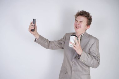 komik gülümseyen genç elinde akıllı telefon ile eğleniyor. beyaz stüdyo arka plan üzerinde kahve kağıt fincan ile ayakta gri iş elbisesi giyen genç çocuk.