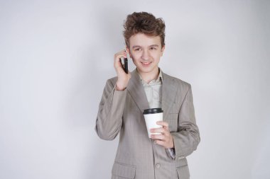 komik gülümseyen genç elinde akıllı telefon ile eğleniyor. beyaz stüdyo arka plan üzerinde kahve kağıt fincan ile ayakta gri iş elbisesi giyen genç çocuk.