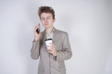 kahve ve akıllı telefon ile kapalı gözleri ile ayakta öğrenci genç çocuk. beyaz stüdyo arka plan üzerinde iş elbisesi giyen çok yorgun genç erkek.