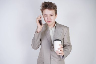 Gri iş elbiseleriyle şaşırmış genç kız cep telefonu ve beyaz stüdyo arka planında bir fincan kahveyle ayakta duruyor.