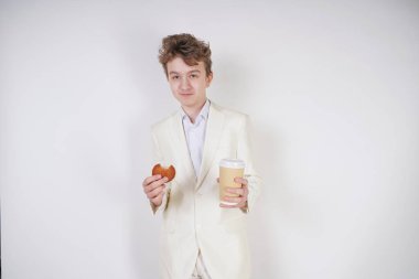 Takım elbiseli genç bir öğrenci elinde bir fincan kahve ve kurabiyeyle stüdyoda beyaz bir arka planda duruyor.