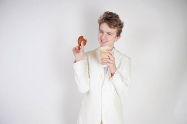 Takım elbiseli genç bir öğrenci elinde bir fincan kahve ve kurabiyeyle stüdyoda beyaz bir arka planda duruyor.