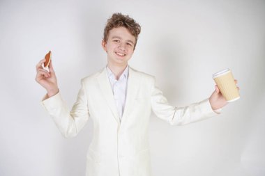 Takım elbiseli genç bir öğrenci elinde bir fincan kahve ve kurabiyeyle stüdyoda beyaz bir arka planda duruyor.