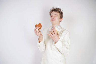 Takım elbiseli genç bir öğrenci elinde bir fincan kahve ve kurabiyeyle stüdyoda beyaz bir arka planda duruyor.