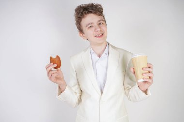 Takım elbiseli genç bir öğrenci elinde bir fincan kahve ve kurabiyeyle stüdyoda beyaz bir arka planda duruyor.
