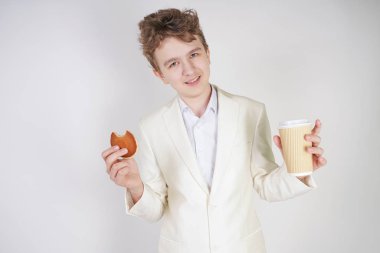 Takım elbiseli genç bir öğrenci elinde bir fincan kahve ve kurabiyeyle stüdyoda beyaz bir arka planda duruyor.