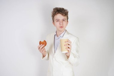 Takım elbiseli genç üzgün öğrenci elinde bir fincan kahve ve kurabiyeyle stüdyoda beyaz bir arka planda duruyor.