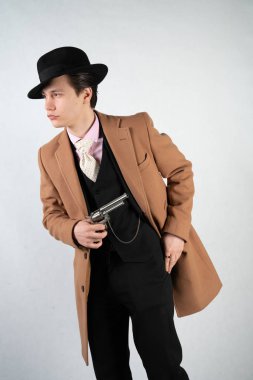 Tek başına stüdyo beyaz bir arka planda elinde bir silah ile resmi bir takım elbise ve şapka genç adam izole