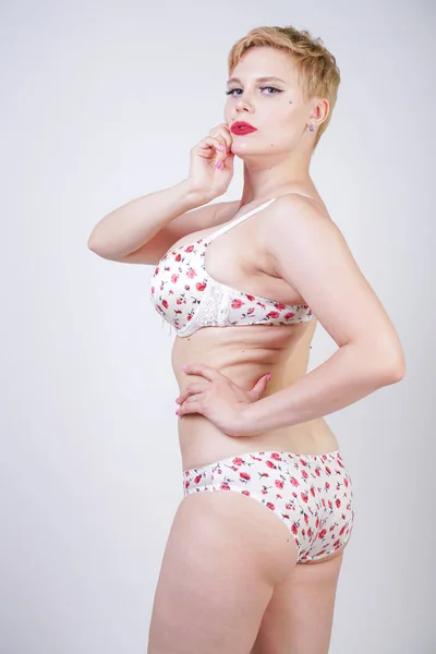 curvy caucasian short hair woman posing in cute white lingerie with red flowers print in the studio alone