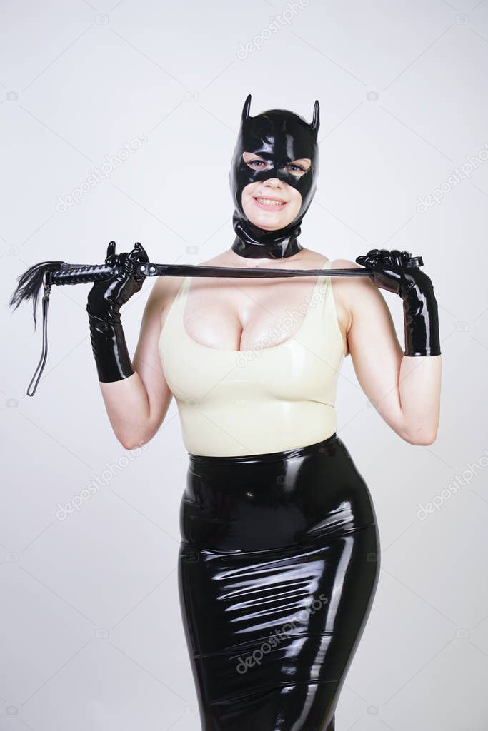 sexy hot latex mistress wearing rubber cat mask and tight black shiny skirt. erotic girl in kinky dress with leather whip on white studio background alone. 