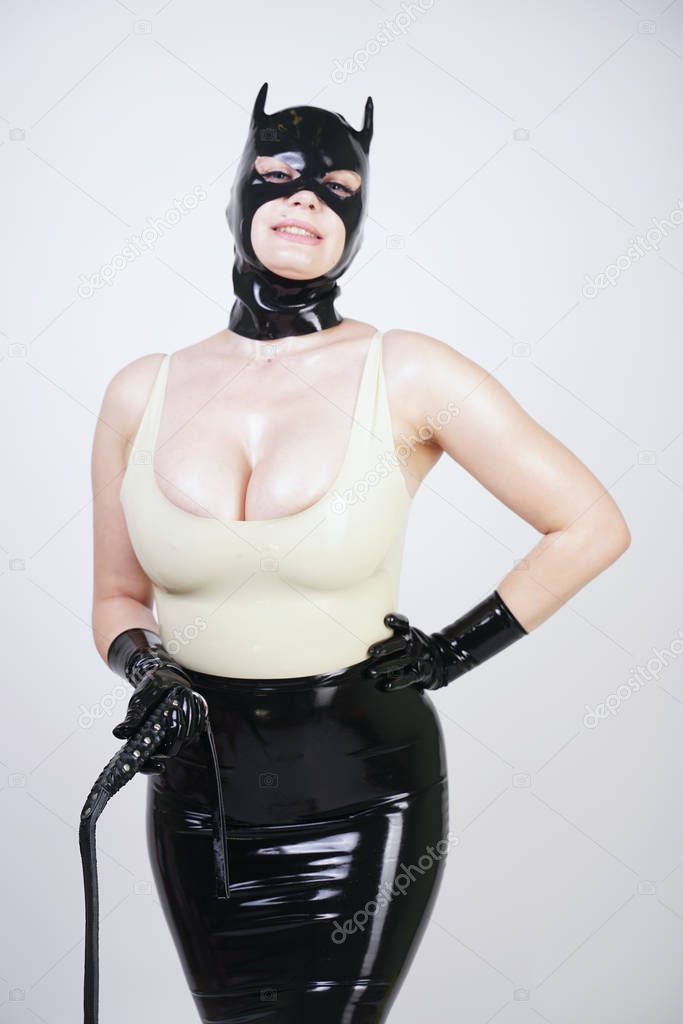 sexy hot latex mistress wearing rubber cat mask and tight black shiny skirt. erotic girl in kinky dress with leather whip on white studio background alone. 