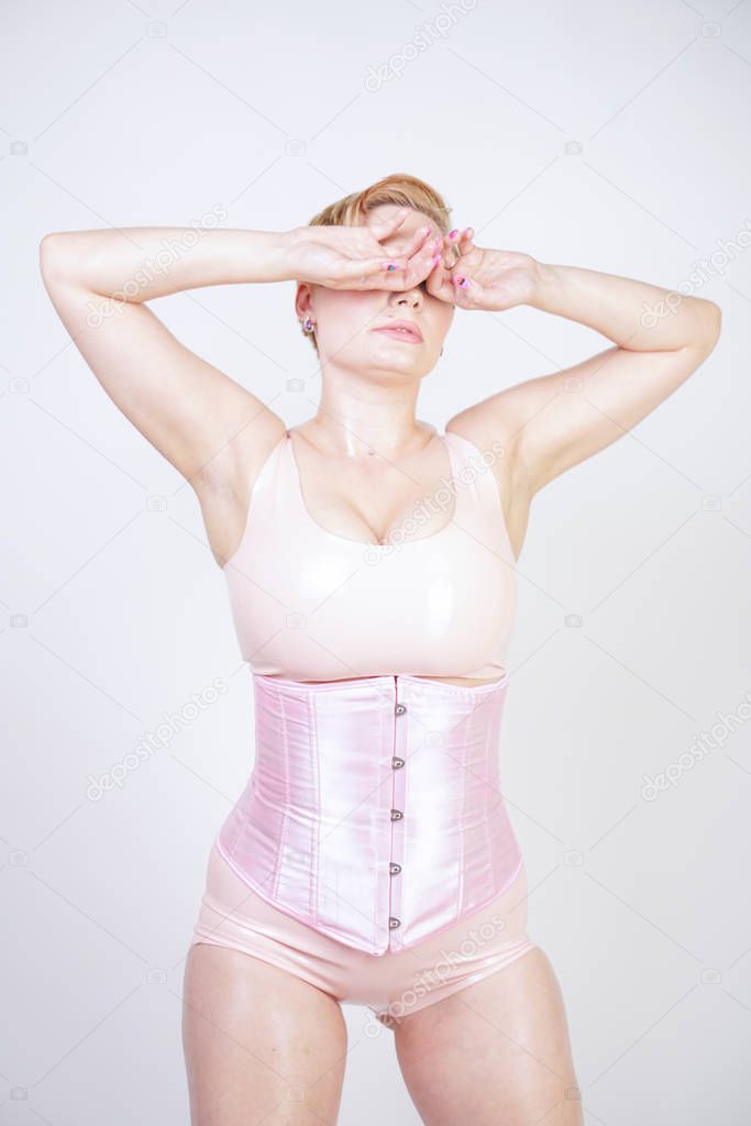 curvy sexy short haired girl in pink latex suit with corset stands on white background in Studio