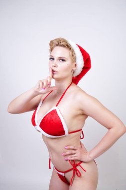 Studio beyaz arka plan üzerinde kırmızı Noel iç çamaşırı kısa saçlı sevimli tombul kız. artı boyutu vücut ile bir kadın standları ve kürk ve bir şapka ile seksi kadife iç çamaşırı pozlar.