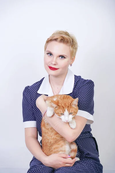 スタジオの白い背景に彼女の最愛のペットを保持ピンナップポルカドットドレスで短い髪を持つかわいい親切な女性 プラスサイズの大人のブロンドの女の子と彼女のオレンジ色の猫は一緒に幸せです — ストック写真