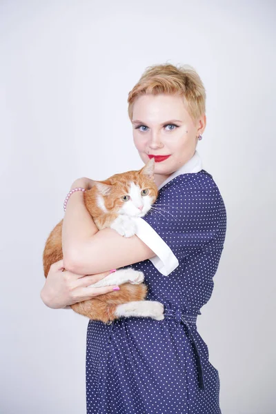 スタジオの白い背景に彼女の最愛のペットを保持ピンナップポルカドットドレスで短い髪を持つかわいい親切な女性 プラスサイズの大人のブロンドの女の子と彼女のオレンジ色の猫は一緒に幸せです — ストック写真